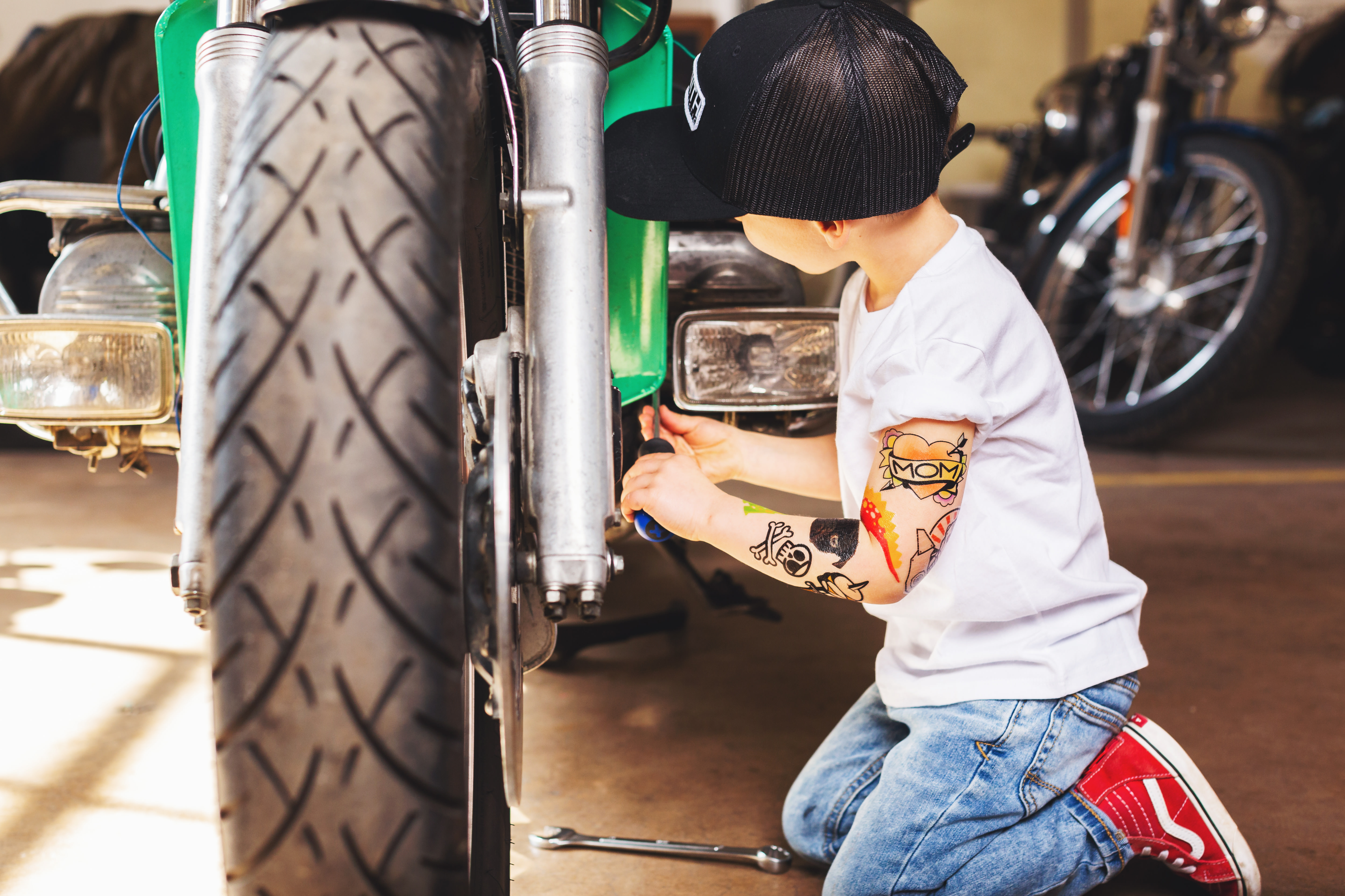 बच्चा बेसबॉल हैट और कस्टम टेम्पररी टैटू पहने हुए मोटरसाइकिल पर काम कर रहा है